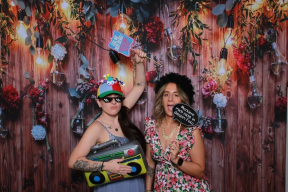Zwei Frauen posieren mit lustigen Requisiten vor der Fotobox und einem Blumen-Hintergrund bei einer Hochzeit in Fürstenfeldbruck.